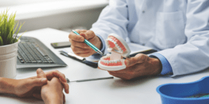 A dentist in a white coat holding a dental model and explaining oral health to a patient during a consultation at a desk with a keyboard, notepad, and dental tools.